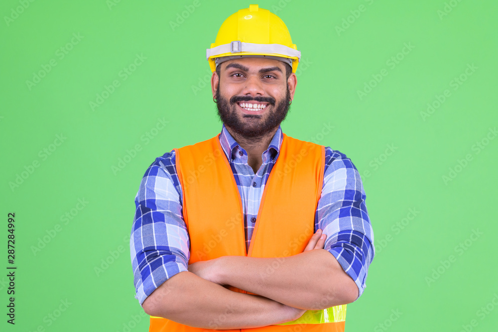 Happy young overweight bearded Indian man construction worker smiling with arms crossed Happy young overweight bearded Indian man construction worker smiling with arms crossed
