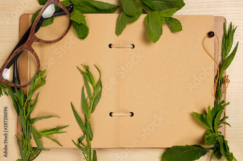 An open notebook of craft paper lies surrounded by herbs with glasses on a wooden table