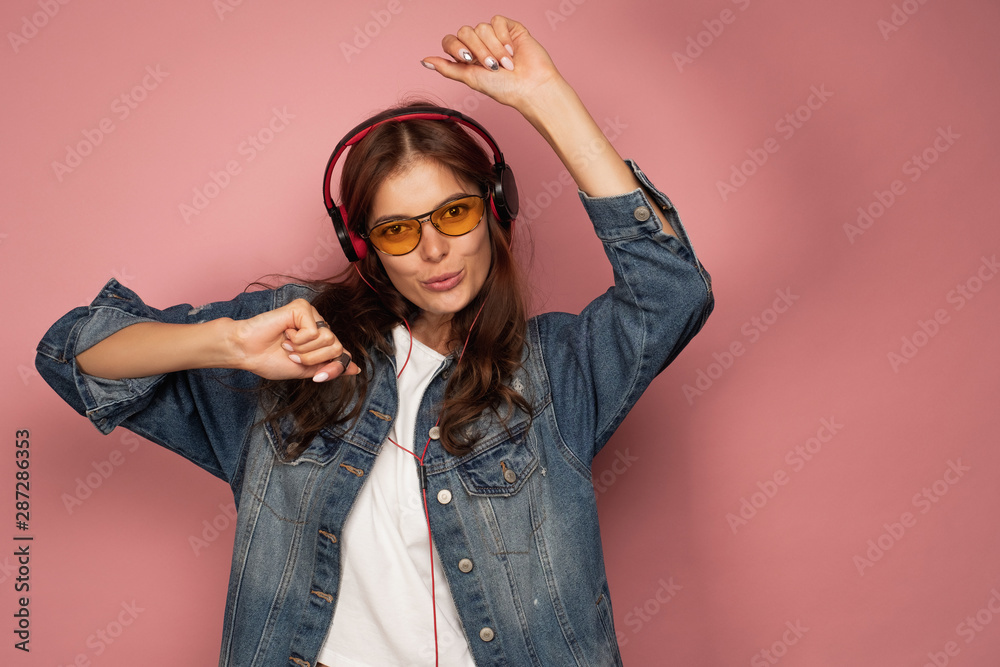 A dark-haired girl in jeans stands on a pink background with headphones, dancing, looking at the camera. A dark-haired girl in jeans stands on a pink background with headphones, dancing, looking at the camera.