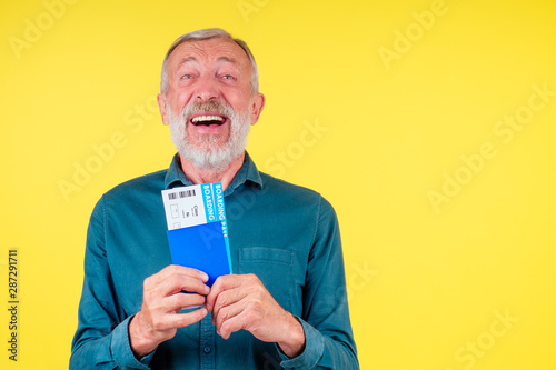 smiling senior man holding passport in blue cover and tickets studio yellow background