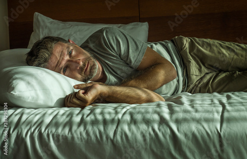 dramatic lifestyle portrait of young attractive sad and depressed man lying on bed at home bedroom thoughtful and pensive feeling overwhelmed and unhappy