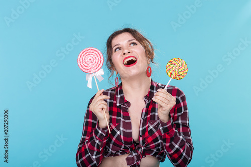 Charming young woman in retro clothes holding colorful lollipops in her hands and licking one posing on blue background. Concept of love for sweets and holidays.