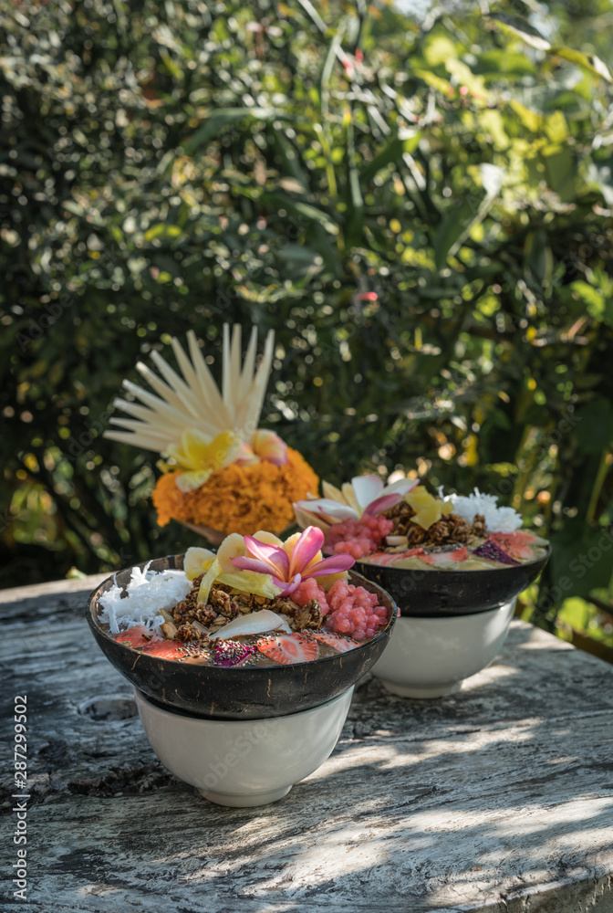 smoothie bowl in a coconut shell plate with with strawberries, chia seeds, banana, granola, flower on wooden background. Green tropical leaves background