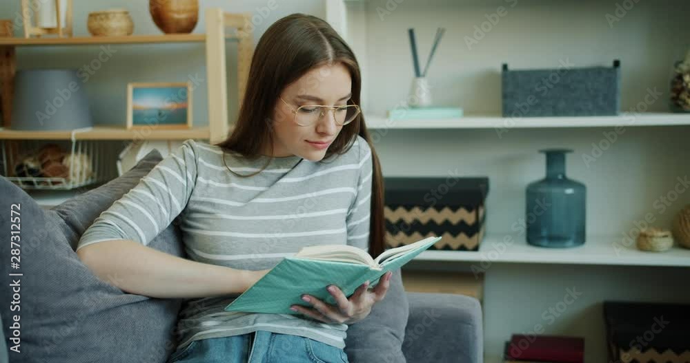 Smiling young woman student is reading interesting book indoors in cozy light studio apartment. Smart lady is wearing casual clothing and trendy glasses. Smiling young woman student is reading interesting book indoors in cozy light studio apartment. Smart lady is wearing casual clothing and trendy glasses.