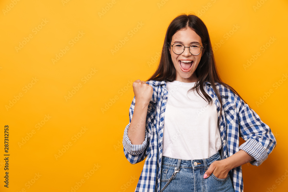 Emotional happy young cute teenage girl in glasses posing isolated over yellow wall background showing winner gesture.