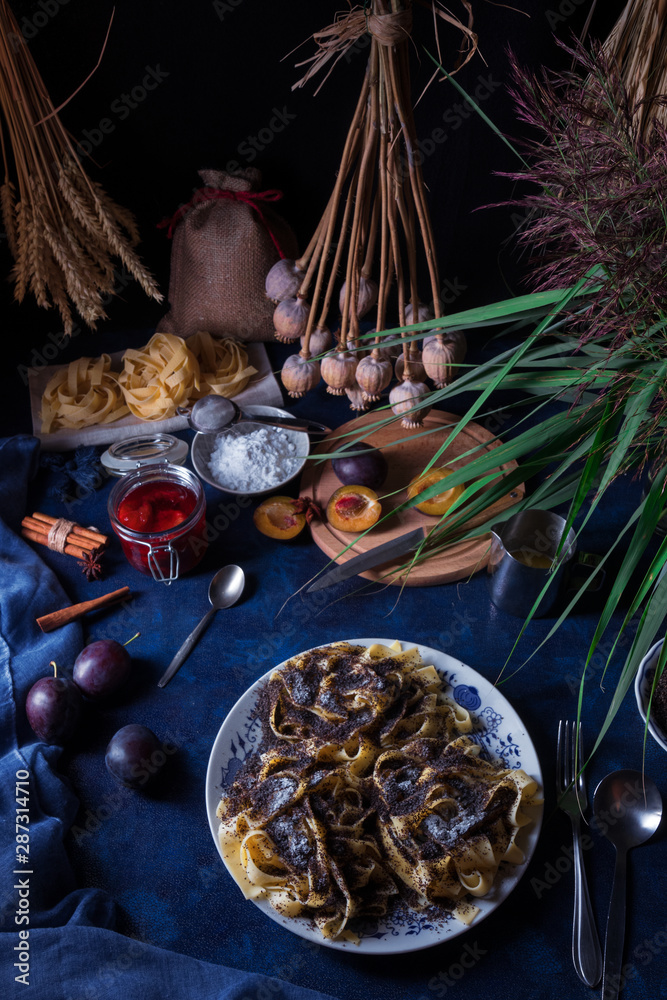 Sweet pasta dessert, noodles with poppy seeds, plum compote from fresh plums, dark background, blue table decorated with fresh flowers. Dry whole poppy plant in background. Sweet pasta dessert, noodles with poppy seeds, plum compote from fresh plums, dark background, blue table decorated with fresh flowers. Dry whole poppy plant in background.
