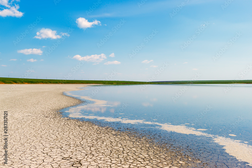 The curved coastline of the salt lake with blue sky, dry cracked soil and tiny stripe of the green grassy land at the horizon. Gruzskoe lake, Rostov-on-Don region, Russia
