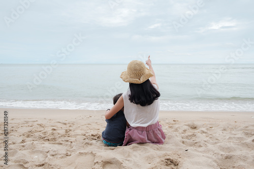 Portrait of  woman sits with her son on the beach and looks at the sea. Back view mom hug her son on beach and point her hand at sky. Mom, lovely concept.