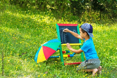 A preschooler boy sits  on the lawn with flowers and draws with chalk on the blackboard.