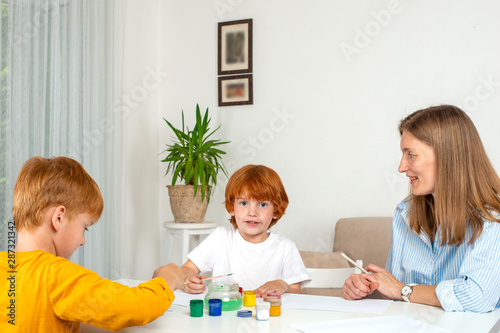 Two little red-haired boys with a nanny or mother or teacher paint with colors.