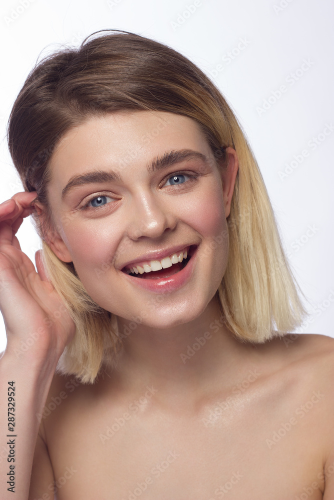 Naklejka premium Closeup of beautiful young woman's face with clean perfect skin and hair smiling at camera. Portrait of a blonde with blue eyes. Spa, skin care and wellness. Isolated on white background