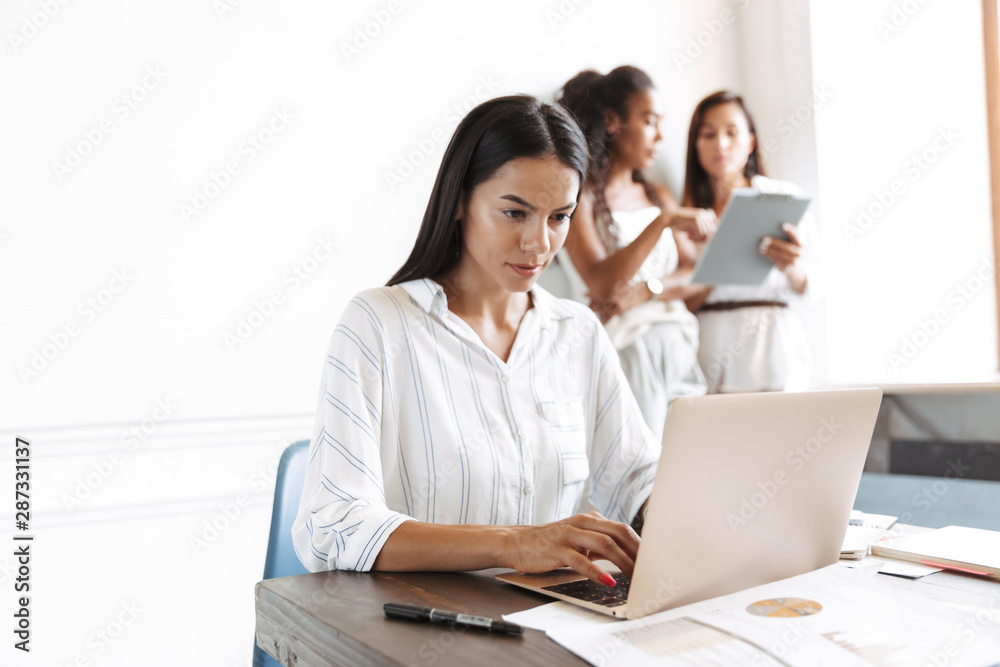Concentrated young business woman at workplace indoors using laptop computer with blurred colleagues on the background. Concentrated young business woman at workplace indoors using laptop computer with blurred colleagues on the background.