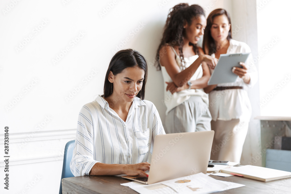 Concentrated young business woman at workplace indoors using laptop computer with blurred colleagues on the background. Concentrated young business woman at workplace indoors using laptop computer with blurred colleagues on the background.