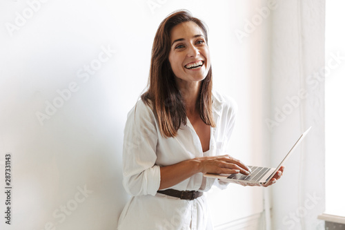 Cheerful smiling young business woman at workplace indoors near window using laptop computer.