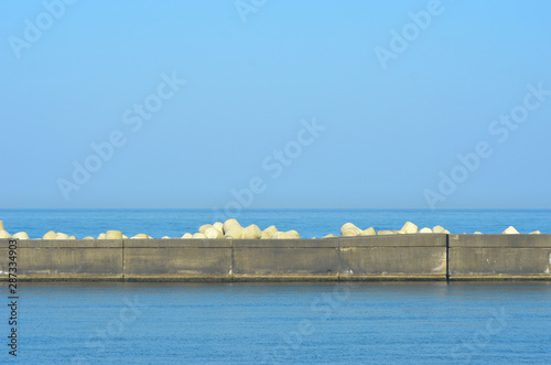 Concrete blocks of a breakwater are seen against calm bue water. The horizon is in the distance  and the sky is blue. White triangular counterweights are on top of the breakwater.