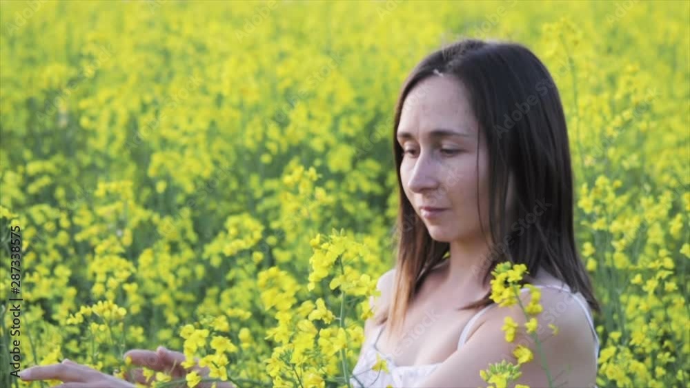 A beautiful carefree girl with healthy hair and freckles on her face against a yellow rapeseed field. Attractive brunette in white dress enjoys yellow rape flowers, natural beauty. A beautiful carefree girl with healthy hair and freckles on her face against a yellow rapeseed field. Attractive brunette in white dress enjoys yellow rape flowers, natural beauty.