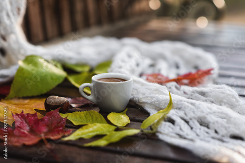 Ceramic cup of coffee with foam, white khitted shawl, dry colorful fallen leaves and chestnut on wet wooden bench in autumn. Close-up photo