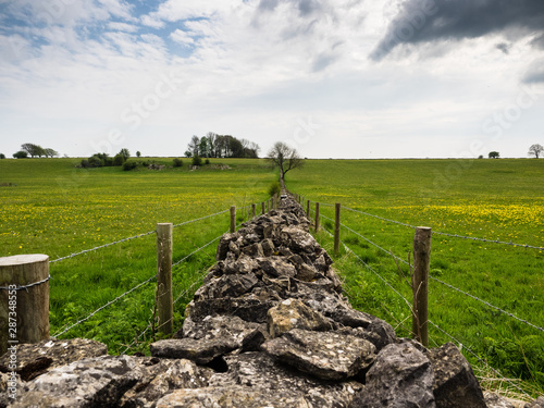 Stone wall in Somerset with barbed wire fence each side and trees and clouds in distance
