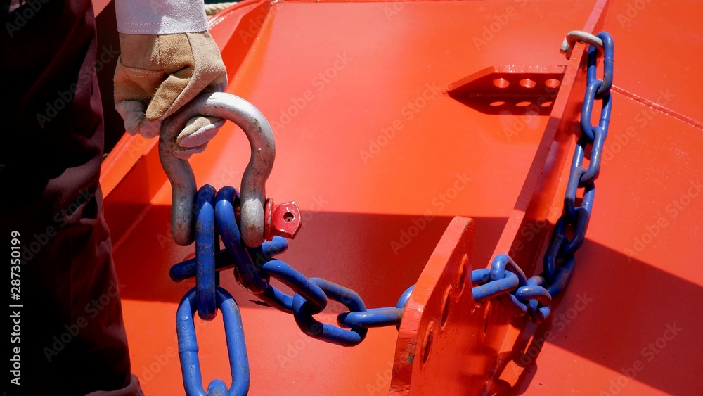 Close up worker hand in leather glove holding bolt type anchor shackle and blue chains with sunlight and shadow on rustproof orange trawl door in trawler fishing vessel Close up worker hand in leather glove holding bolt type anchor shackle and blue chains with sunlight and shadow on rustproof orange trawl door in trawler fishing vessel