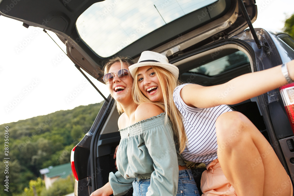 Two female best friends on travel. They're sitting on a car trunk and relaxing after a long journey.