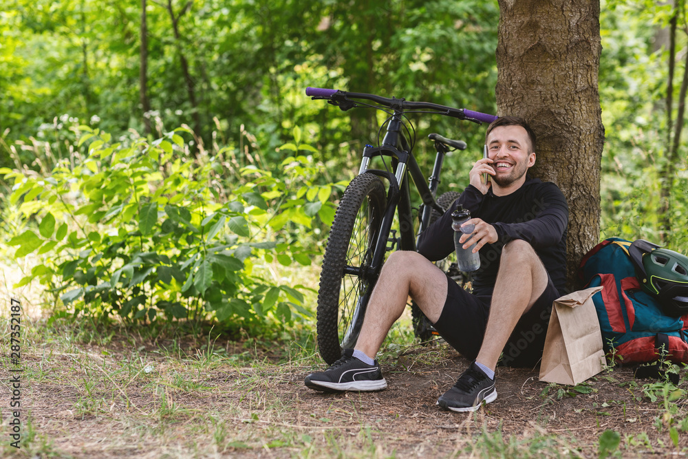 Male cyclist resting under tree after exercising, talking by phone