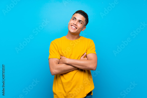 Young man with yellow shirt over isolated blue background looking up while smiling