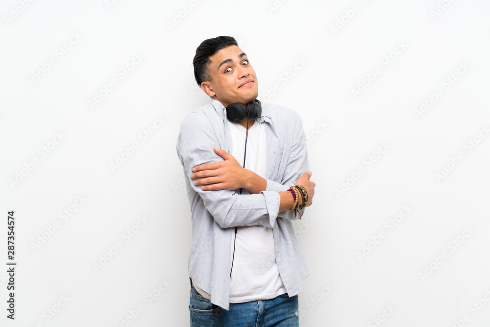 Young man over isolated white wall with earphones making doubts gesture while lifting the shoulders Young man over isolated white wall with earphones making doubts gesture while lifting the shoulders