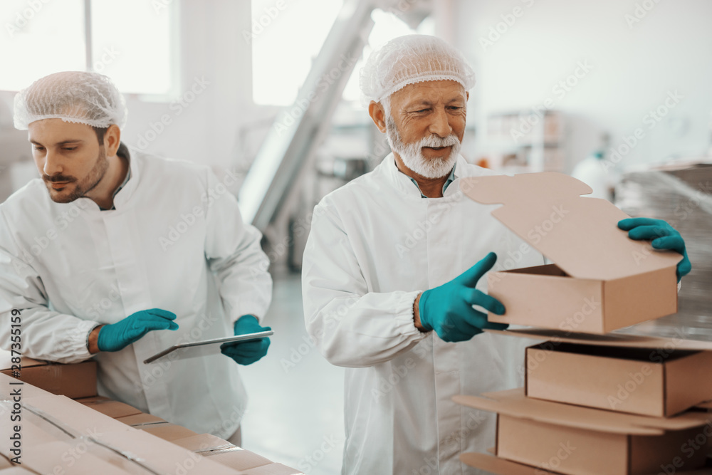 Caucasian working senior in white uniform putting and preparing boxes while smiling supervisor holding tablet and checking boxes. Food plant interior. Caucasian working senior in white uniform putting and preparing boxes while smiling supervisor holding tablet and checking boxes. Food plant interior.