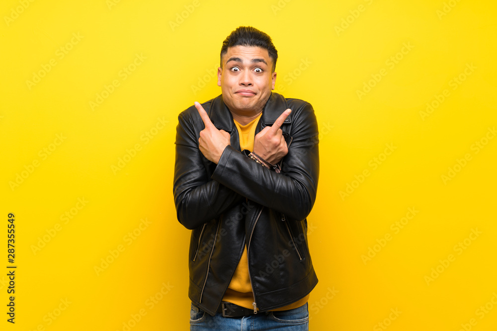 Young man over isolated yellow background pointing to the laterals having doubts Young man over isolated yellow background pointing to the laterals having doubts