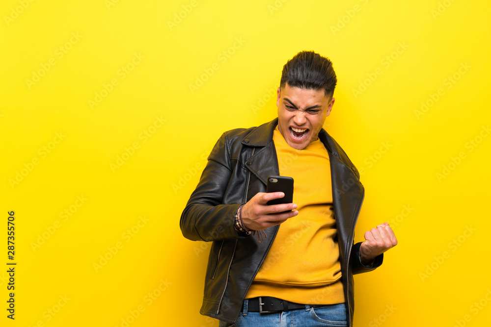Young man over isolated yellow background with phone in victory position Young man over isolated yellow background with phone in victory position
