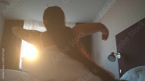 Young woman with long hair wakes up, gets up on bed and stretches in morning sun light
