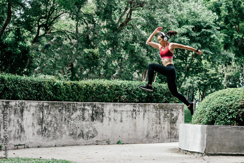 Full-length portrait of young woman trail runner jumping while having workout in city park. Horizontal shot. Side view
