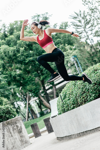 Full-length portrait of young woman trail runner jumping while having workout in city park. Vertical shot. Dutch angle