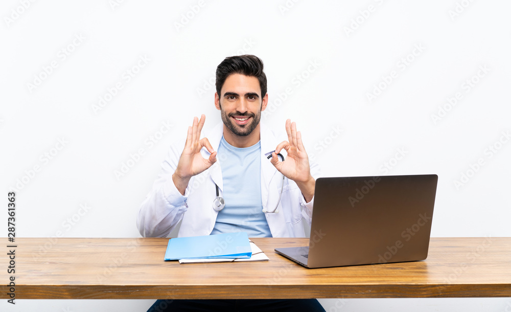 Young doctor man with his laptop over isolated wall showing an ok sign with fingers Young doctor man with his laptop over isolated wall showing an ok sign with fingers