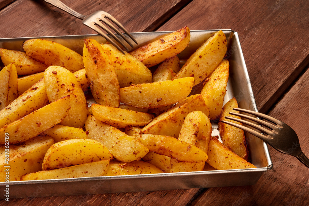 Potato wedges, oven baked, in a baking tray on a dark rustic wooden background, with two forks Potato wedges, oven baked, in a baking tray on a dark rustic wooden background, with two forks
