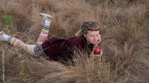 Girl laying on the wild grass and eating red apple, enjoying summer morning . Summer lifestyle portrait of trendy hipster girl with dreads. photo