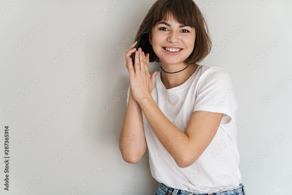 Smiling positive amazing young lady isolated over grey wall background talking by mobile phone covering microphone. Smiling positive amazing young lady isolated over grey wall background talking by mobile phone covering microphone.