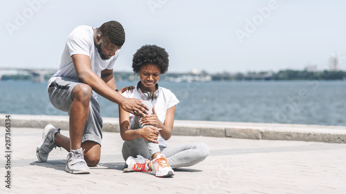 Fototapeta Naklejka Na Ścianę i Meble -  Black Guy Helping Girl Examining Sprained Ankle At Riverbank