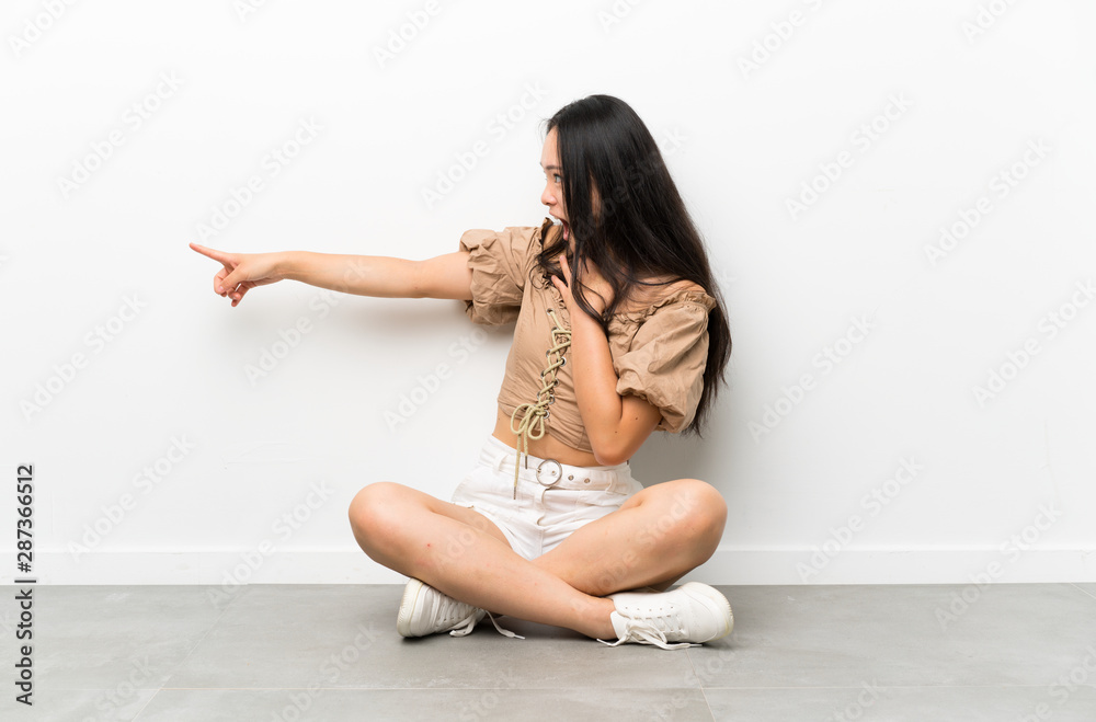 Teenager asian girl sitting on the floor pointing finger to the side Teenager asian girl sitting on the floor pointing finger to the side