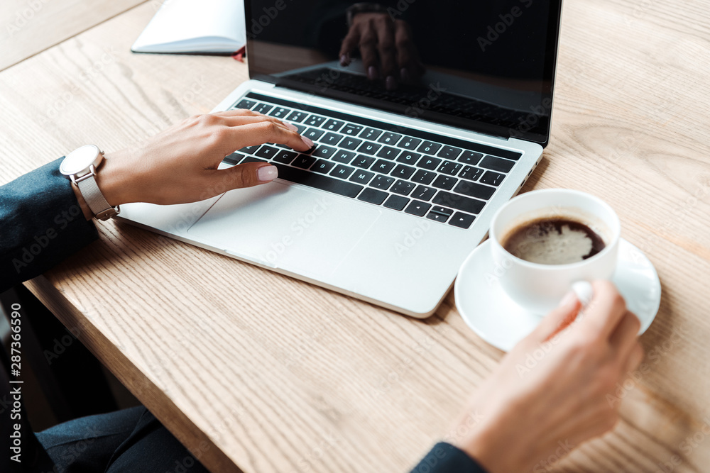 cropped view of businesswoman typing on laptop with blank screen near cup of coffee on table cropped view of businesswoman typing on laptop with blank screen near cup of coffee on table