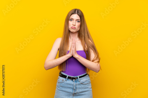 Young woman over isolated yellow background keeps palm together. Person asks for something