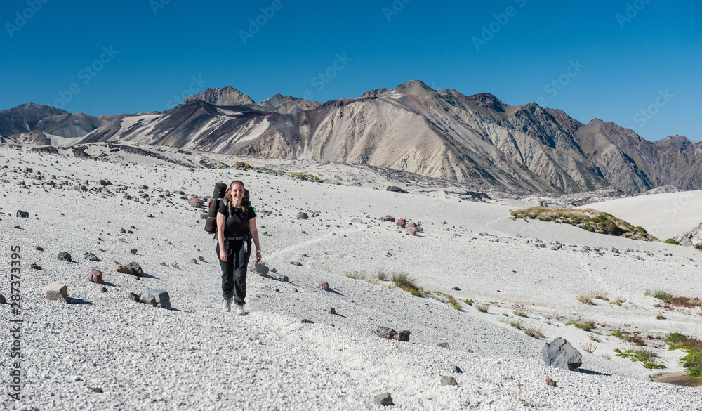 A young woman with backpack in unique white sand desert mountains in Patagonia, Chile. Sunny sky, active volcanos, white mountains. Backpacking, hiking and camping on Condor Circuit in South America.
