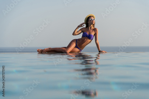 Summer. Woman model in fashion swimsuit lying on edge of infinity swimming pool with sea view.