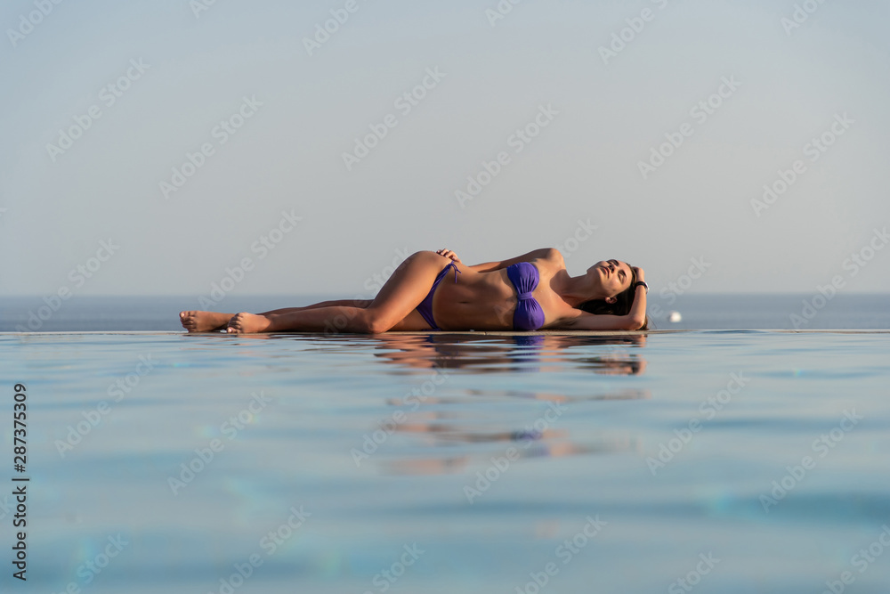 Summer. Woman model in fashion swimsuit lying on edge of infinity swimming pool with sea view.