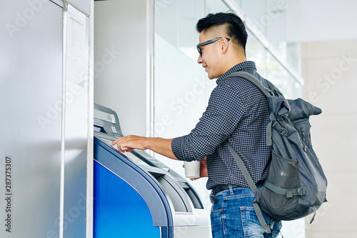 Smiling university student using ATM cash terminal to withdraw money from the credit card