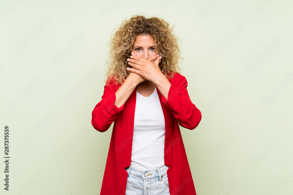 Young blonde woman with curly hair over isolated green background covering mouth with hands Young blonde woman with curly hair over isolated green background covering mouth with hands