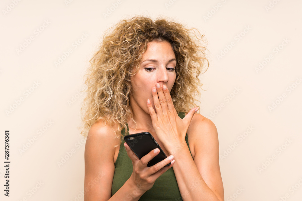 Young blonde woman with curly hair using mobile phone with surprise facial expression