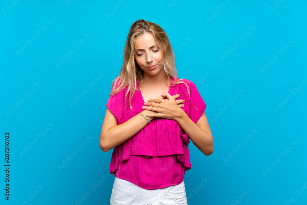 Blonde young woman over isolated blue background having a pain in the heart