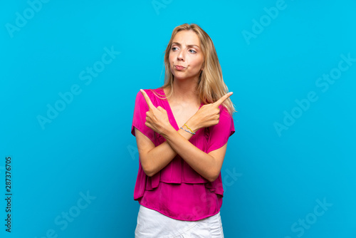 Blonde young woman over isolated blue background pointing to the laterals having doubts © luismolinero