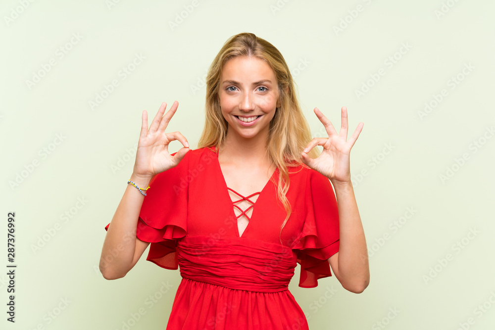 Blonde young woman over isolated green background showing an ok sign with fingers Blonde young woman over isolated green background showing an ok sign with fingers